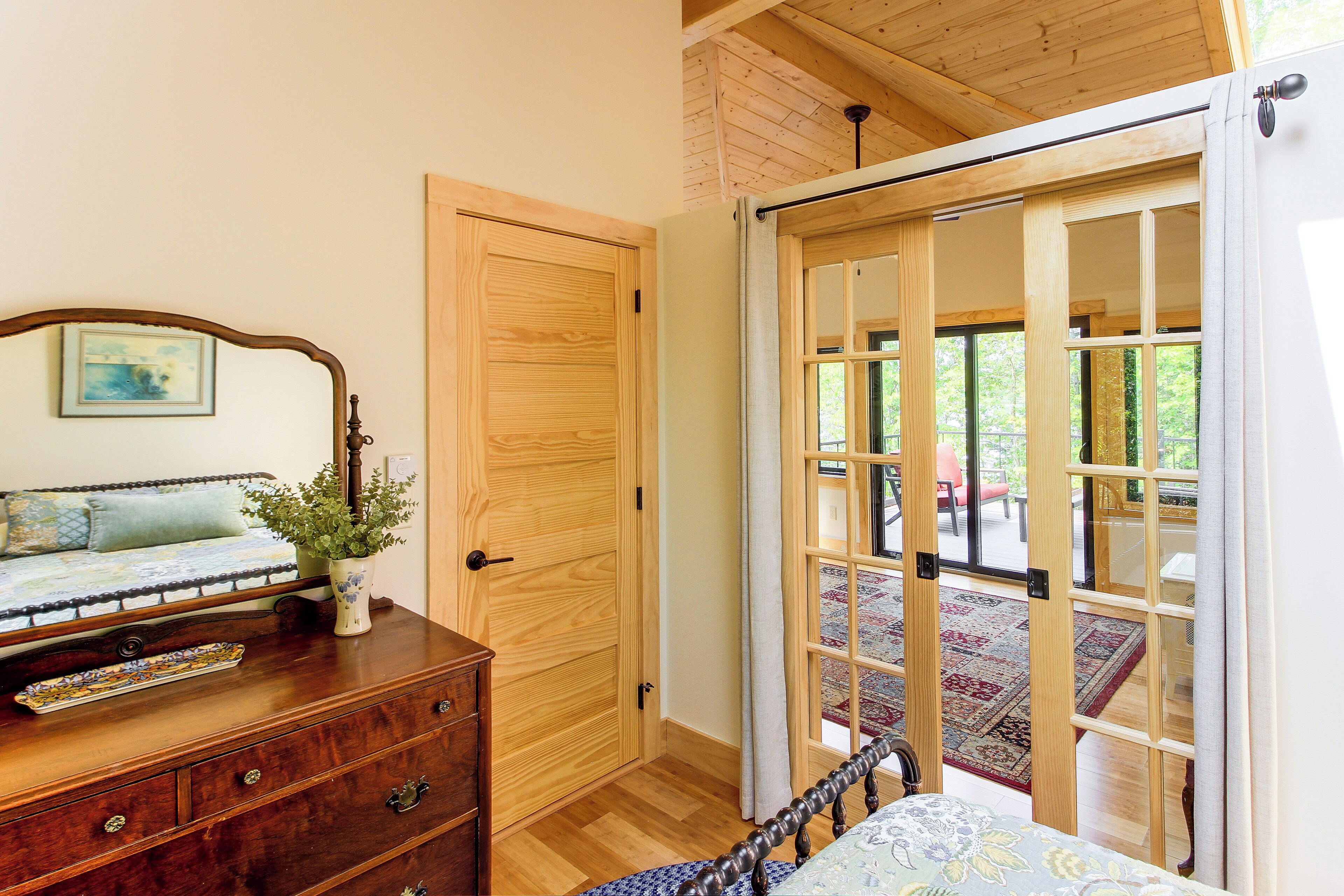 A bedroom with a wooden dresser, a bed with a floral bedspread, a wooden door, and a view of an outdoor deck through a glass door.