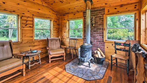 A log cabin interior with a wood-burning stove, wooden furniture, and large windows. - Serene Wilderness Acres Retreat on Bear Lake (Mc Grath)