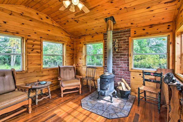 A log cabin interior with a wood-burning stove, wooden furniture, and large windows. - Serene Wilderness Acres Retreat on Bear Lake (Mc Grath)