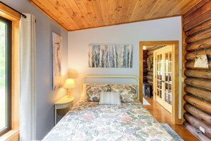 A bedroom with a bed, a nightstand, a lamp, a painting, and a wooden log wall. - Serene Wilderness Acres Retreat on Bear Lake (Mc Grath)