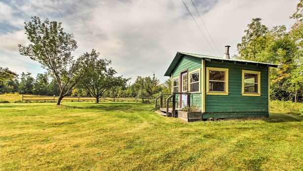Una casita verde con porche y chimenea, rodeada de un campo cubierto de césped y árboles.