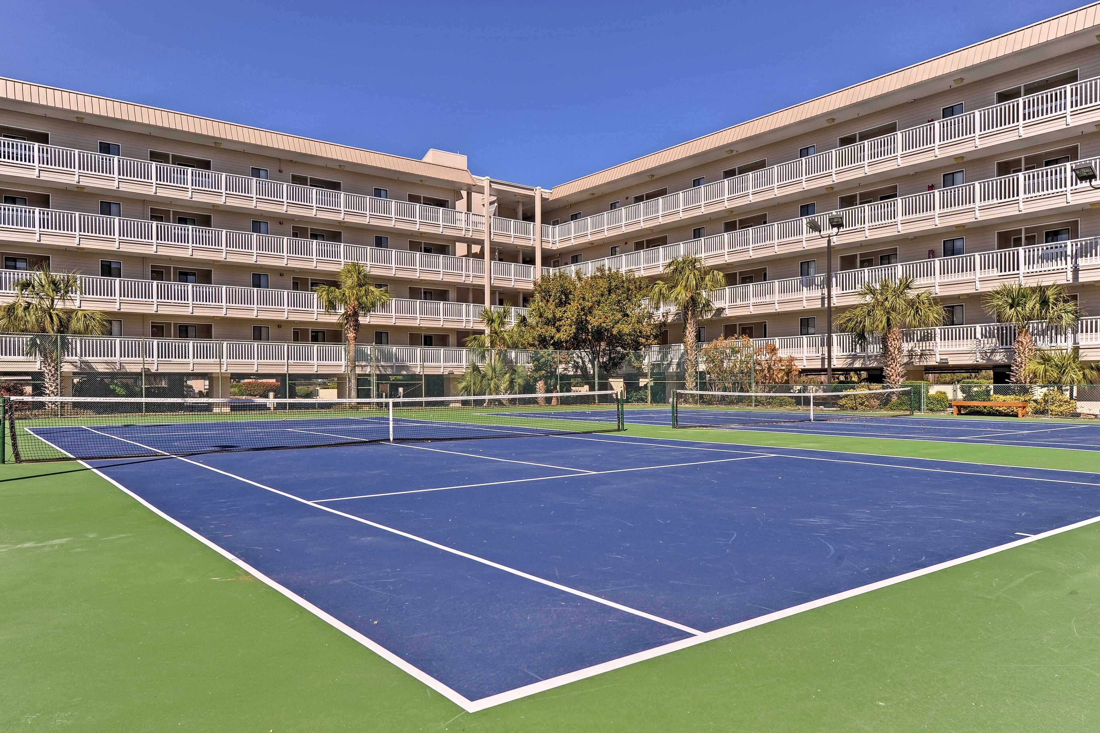 Un court de tennis entouré d’un hôtel à plusieurs étages, doté de balcons et de palmiers.