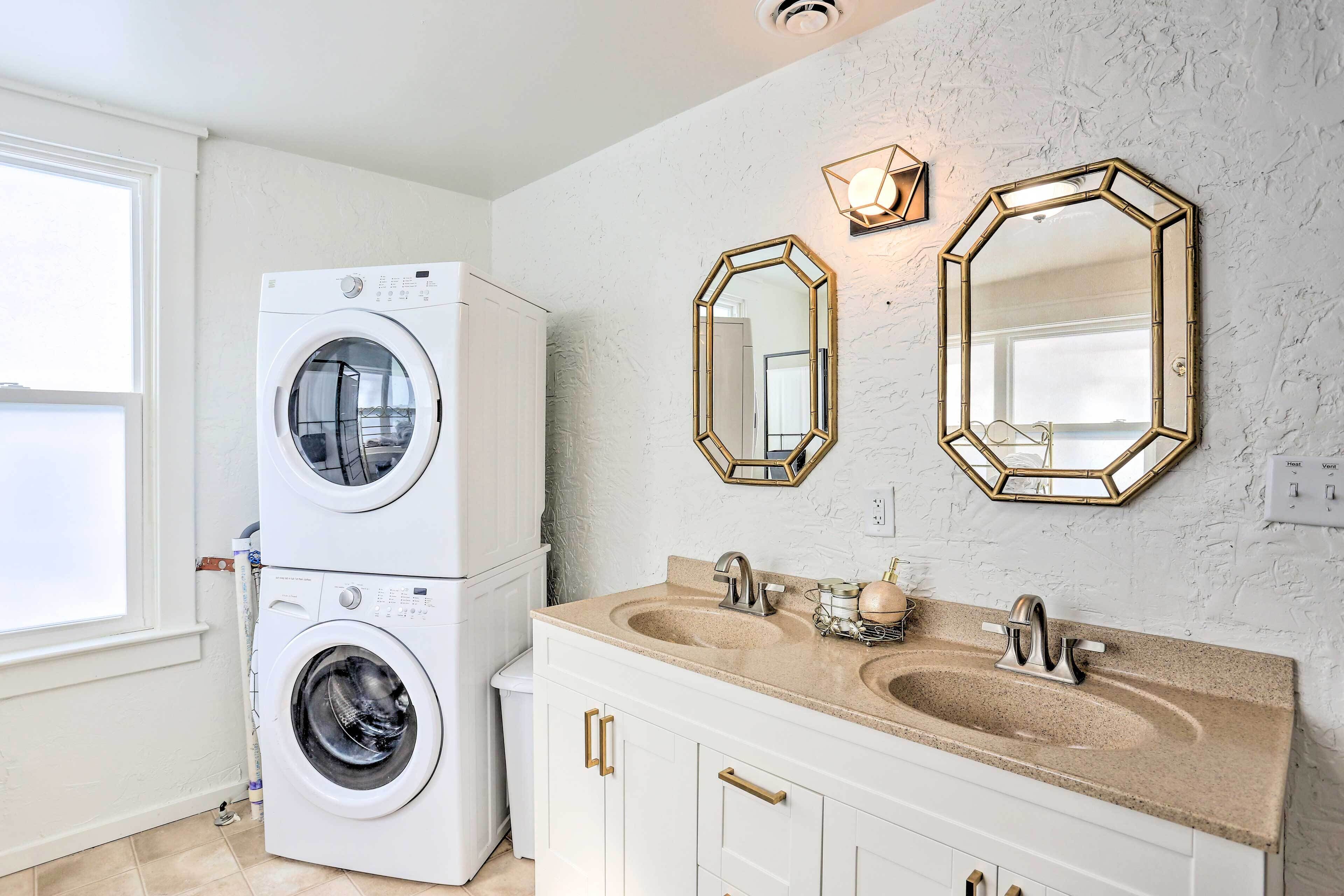 Une petite salle de bain équipée d’un lave-linge et d’un sèche-linge superposés, de deux miroirs fixés au mur, d’un meuble-lavabo à deux vasques et d’un interrupteur pour contrôler le chauffage et la ventilation.