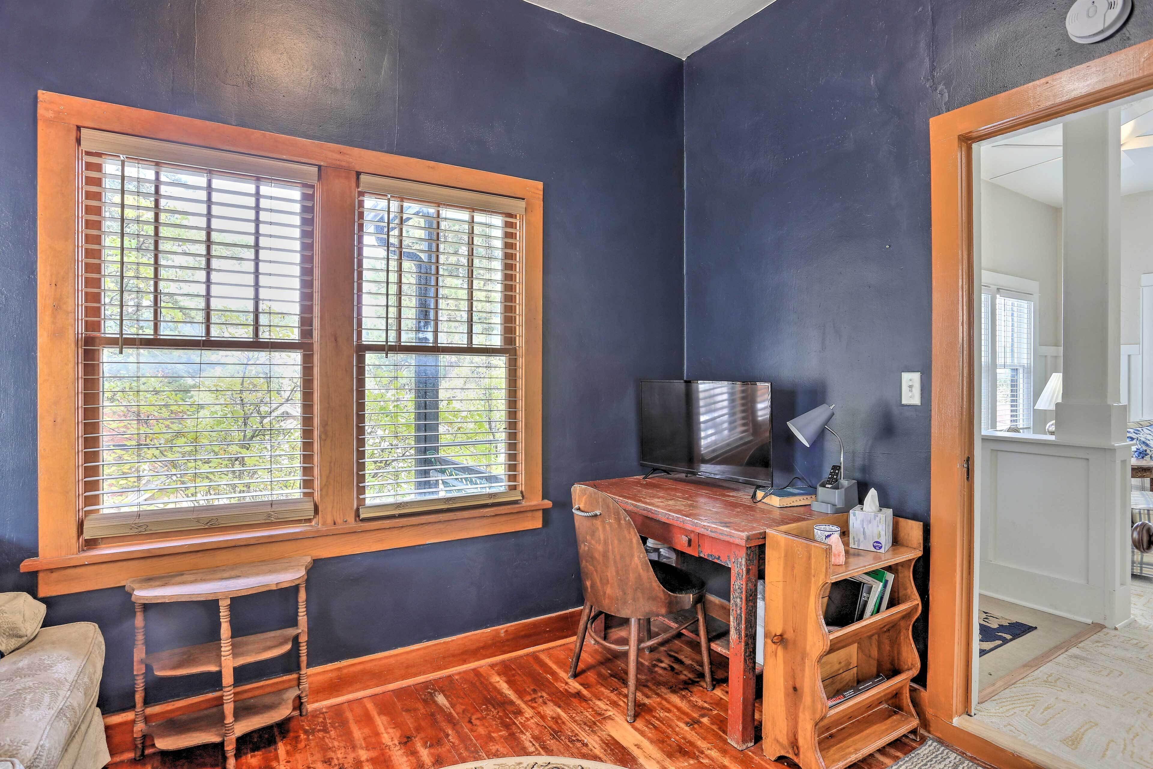 A room with a wooden desk, a chair, and a television. There is a window with blinds and a door leading to another room.
