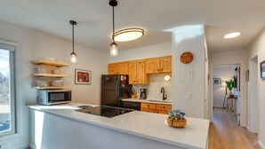 A modern kitchen with white cabinetry, a black refrigerator, a microwave, and a large island with a sink. There are wooden cabinets above the stove and a hanging light fixture. - Walk to Winter Park Bus From Fraser Condo! (Fraser)