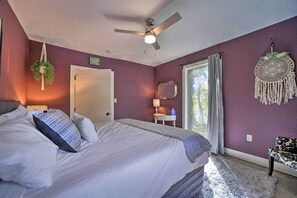 Une chambre à coucher comprenant un lit, un ventilateur de plafond, une fenêtre avec des rideaux, une table de chevet avec une lampe et un élément décoratif mural.