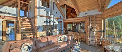 A spacious log cabin interior with a large wooden staircase, a leather sofa with decorative pillows, a dining area, and a kitchen with wooden cabinets.
