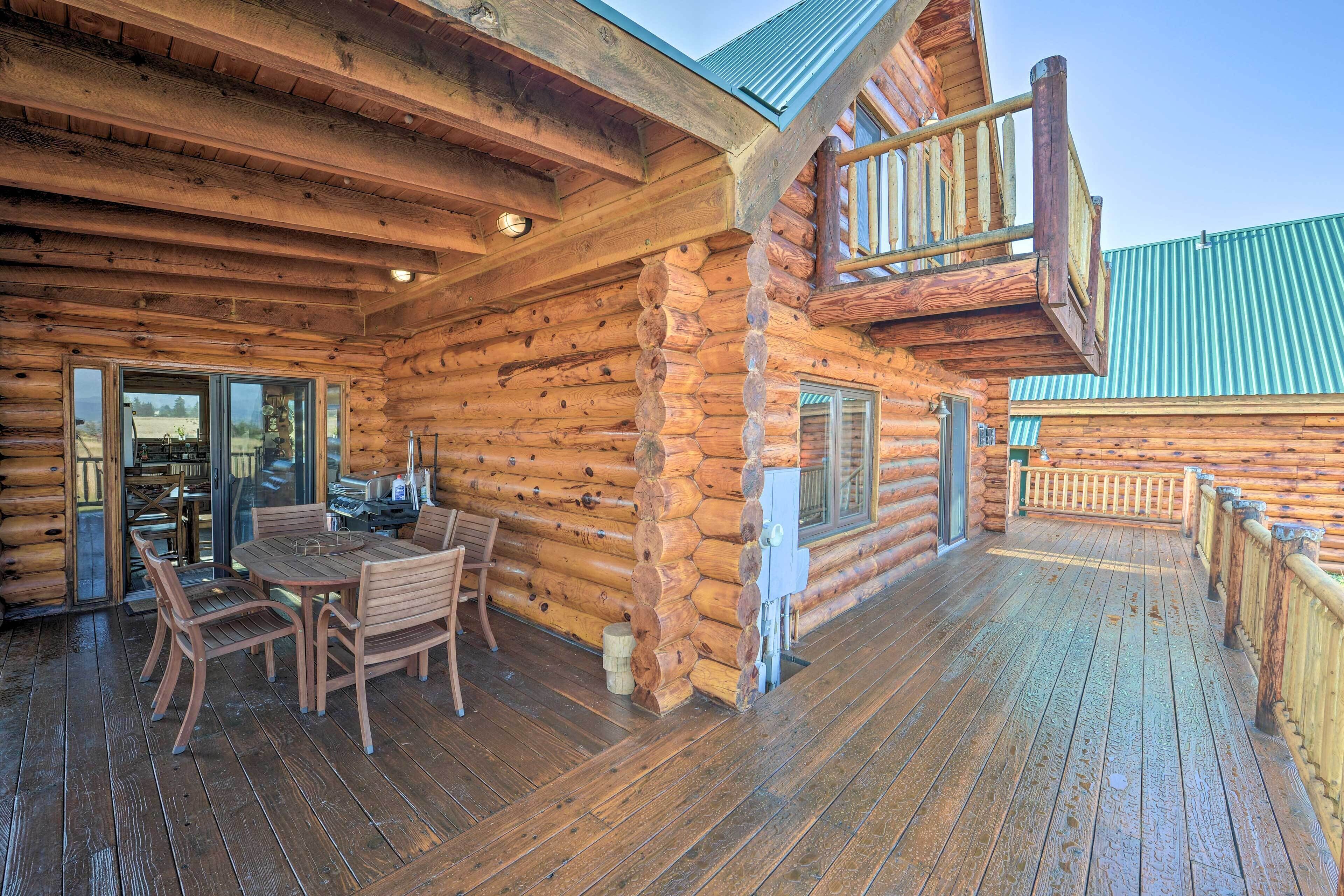 Una cabaña de troncos con una terraza de madera, una zona de comedor con sillas y una mesa, y un balcón con barandilla.