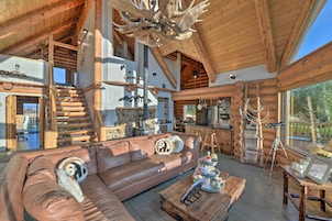 A spacious log cabin interior with a large wooden staircase, a leather sofa with decorative pillows, a dining area, and a kitchen with wooden cabinets.