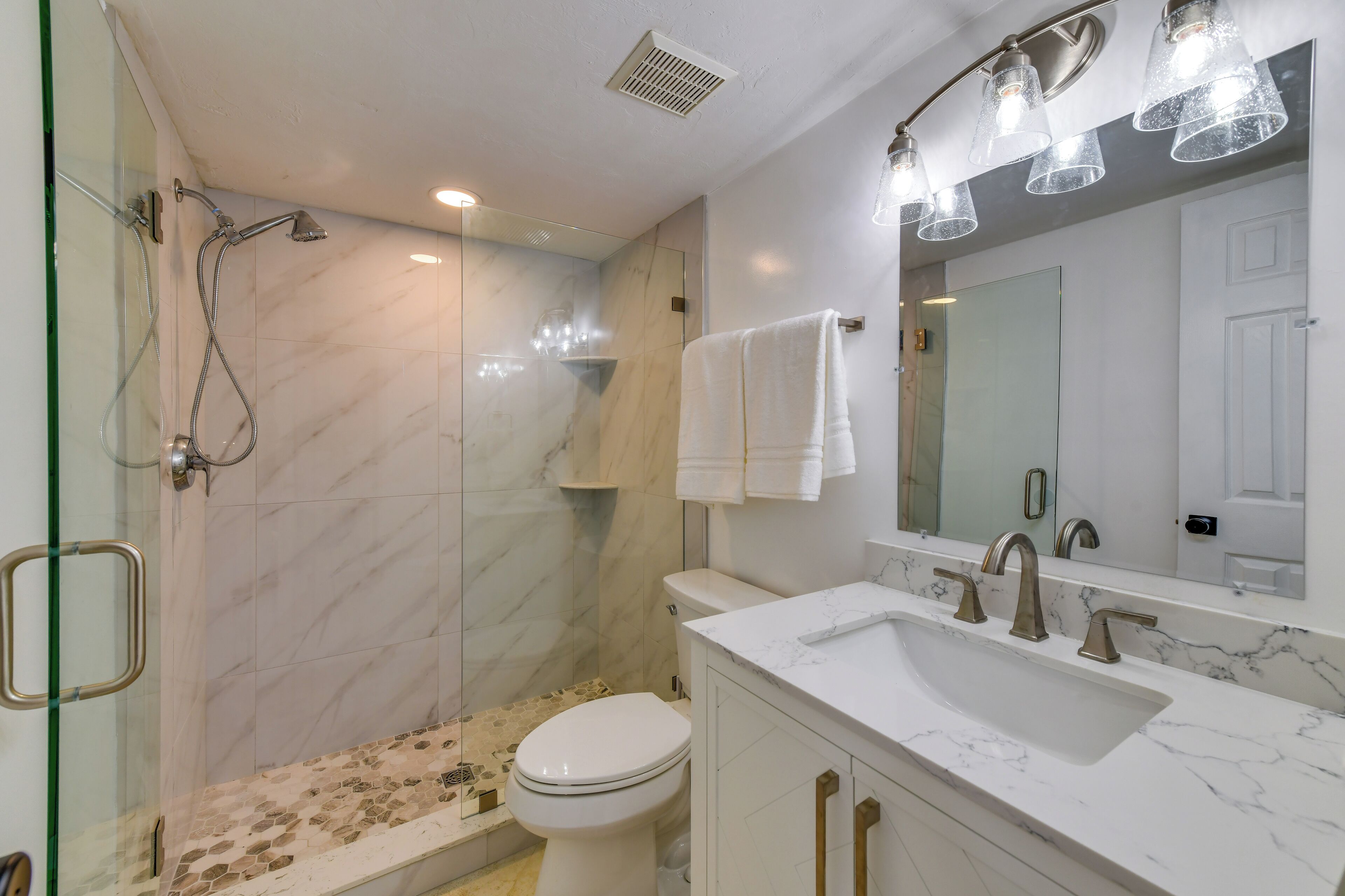 Een badkamer met een douchecabine van glas, een toilet, een wastafel met een marmeren blad en een spiegel met verlichting.