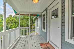 A covered porch with a wooden railing, a welcome mat, and a view of a road and greenery.