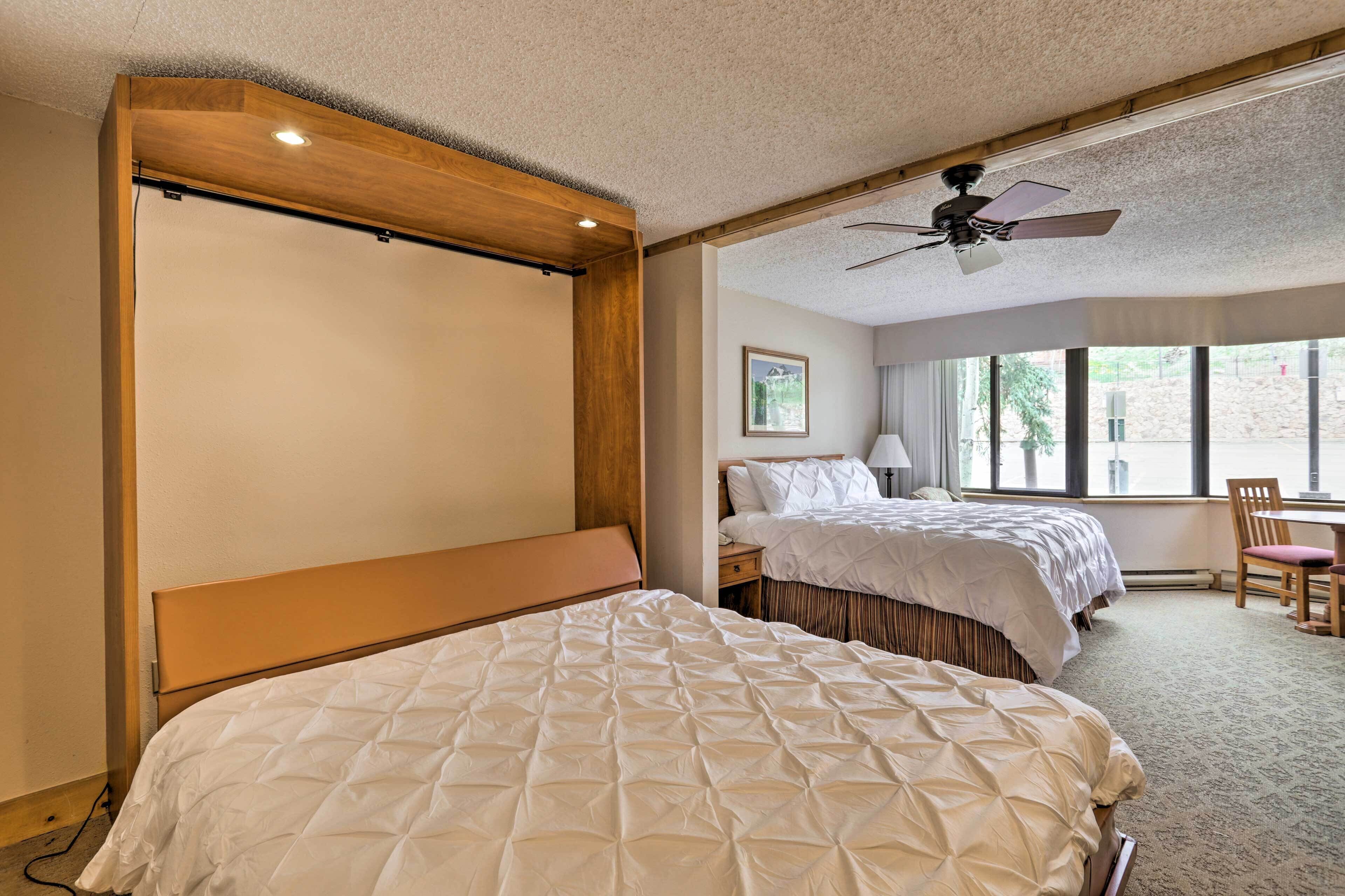 Habitación de hotel con dos camas, ventilador de techo, escritorio con silla y ventana con vistas.