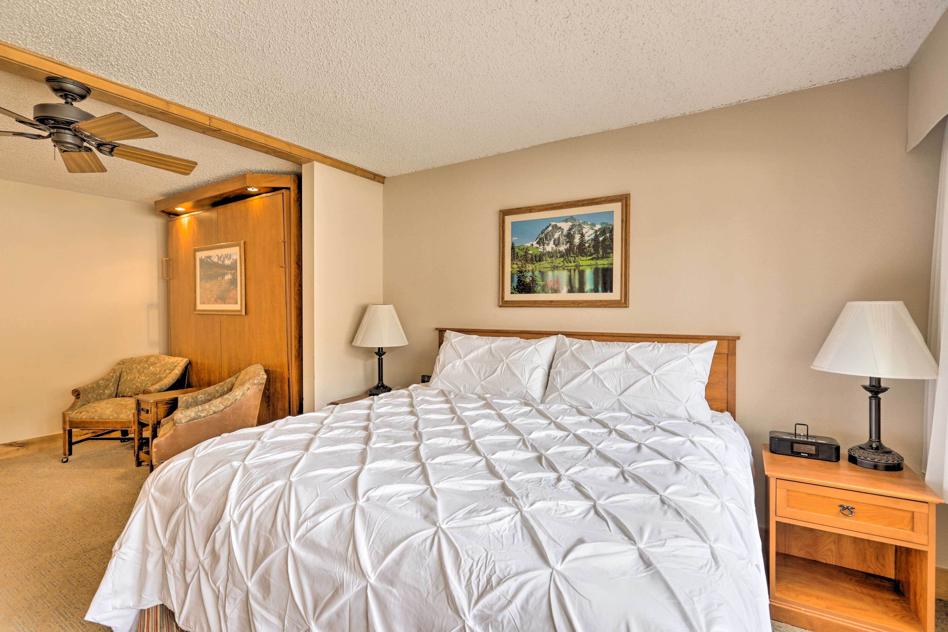 A bedroom with a large bed, a wooden nightstand, a chair, a ceiling fan, and a painting on the wall.
