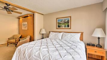 A bedroom with a large bed, a wooden nightstand, a chair, a ceiling fan, and a painting on the wall.