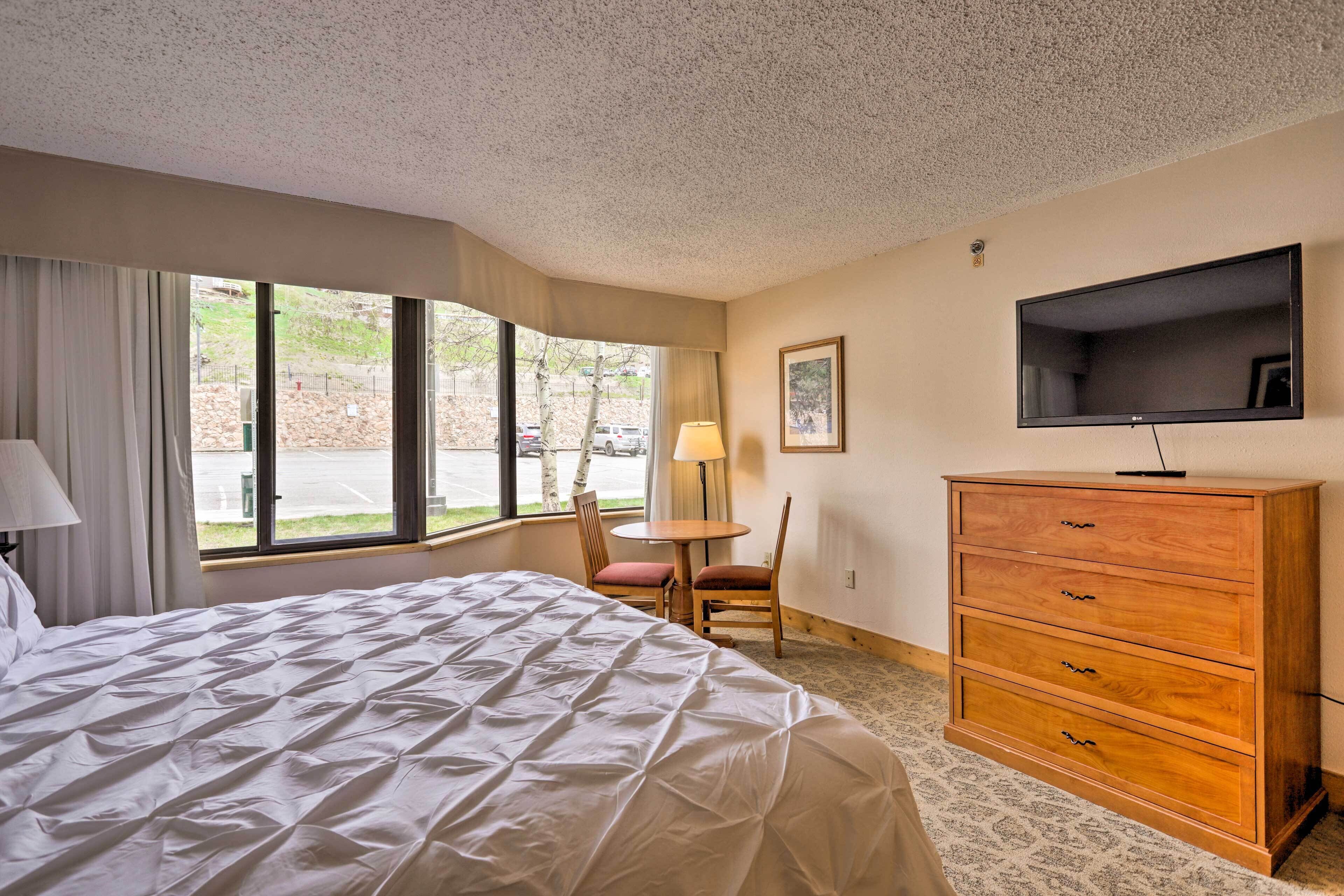 A hotel room with a large bed, a wooden dresser, a television, a window with curtains, and a small table with chairs.