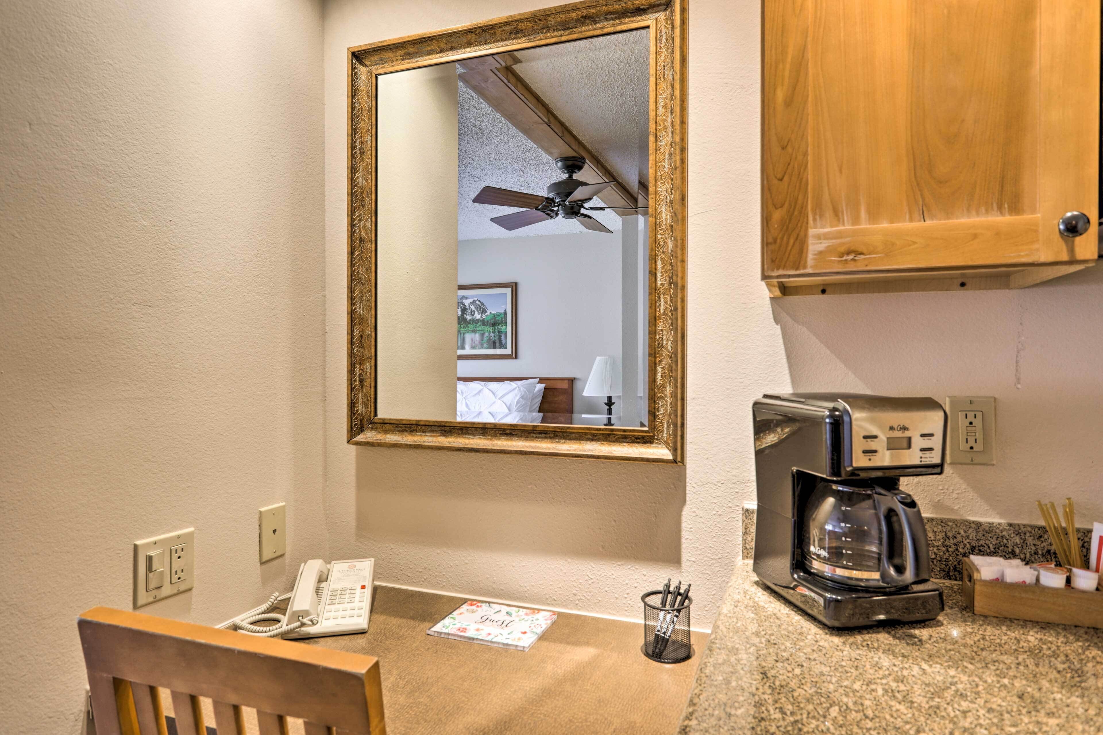 Une chambre d’hôtel avec une chaise en bois, un bureau avec un téléphone, une machine à café et un miroir qui reflète un ventilateur de plafond et un lit avec un oreiller blanc.