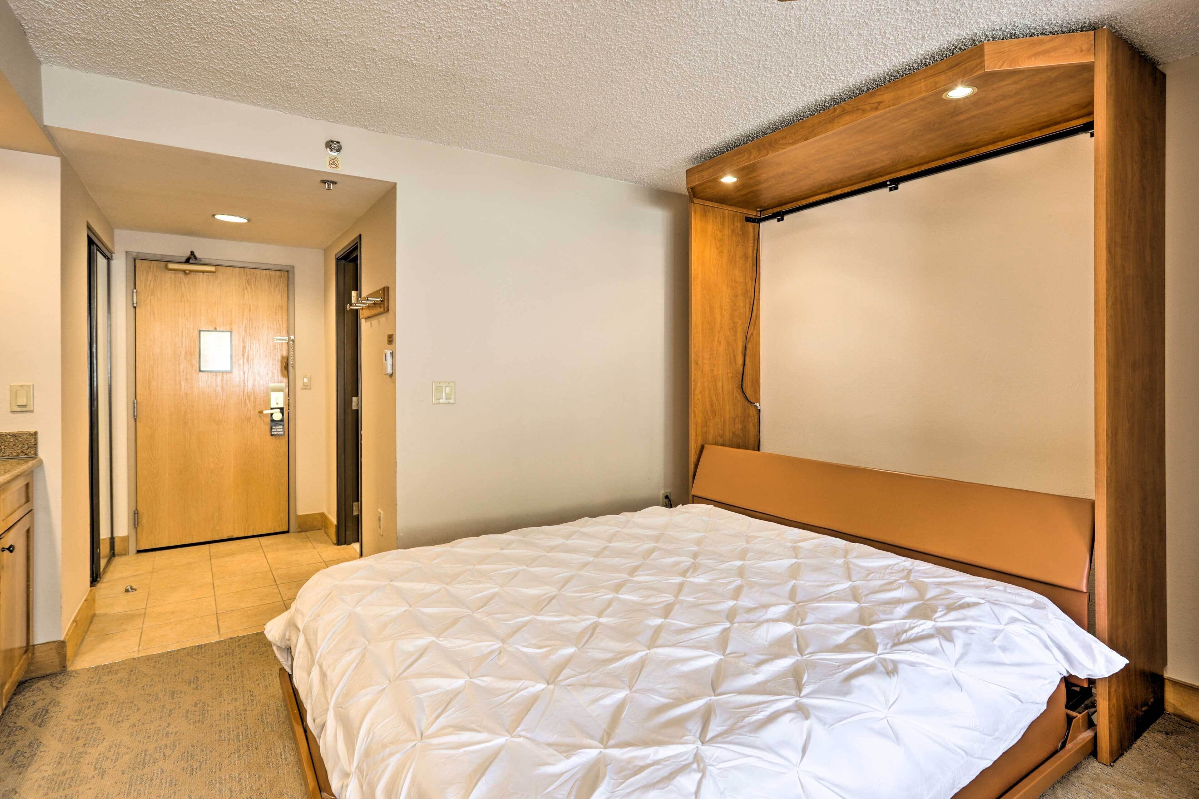 A hotel room with a large bed, wooden headboard, and a door leading to a hallway.