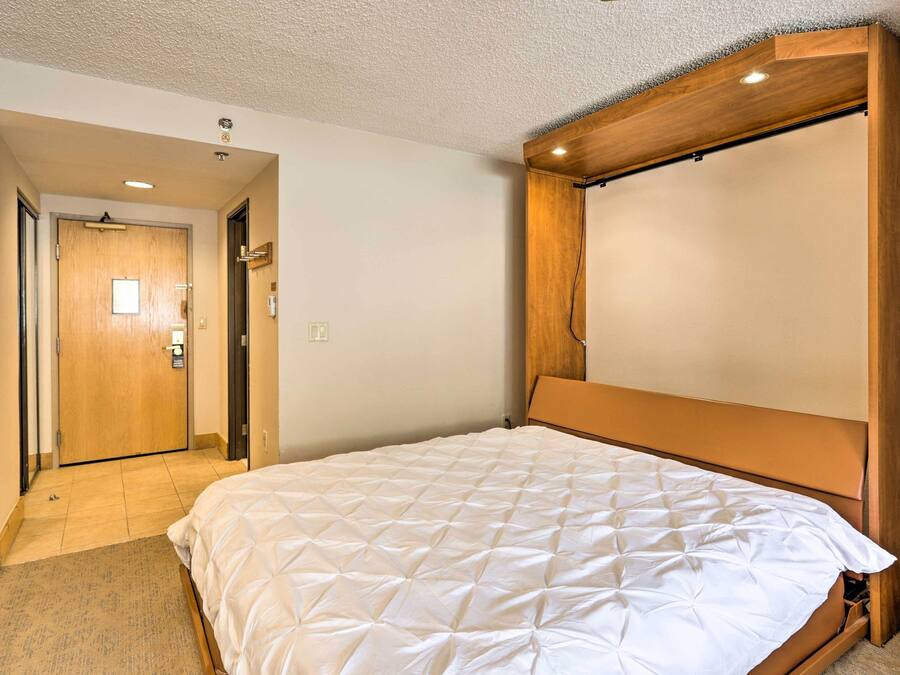 Quarto de hotel com uma cama grande, cabeceira de madeira e porta que dá para um corredor.