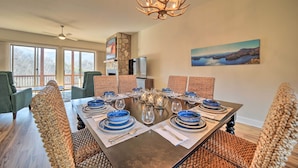 A dining area with a long table set for six, surrounded by woven chairs, a chandelier, and a stone accent wall. - Beautiful Whittier Condo w/ Deck + Mtn Views! (Whittier)