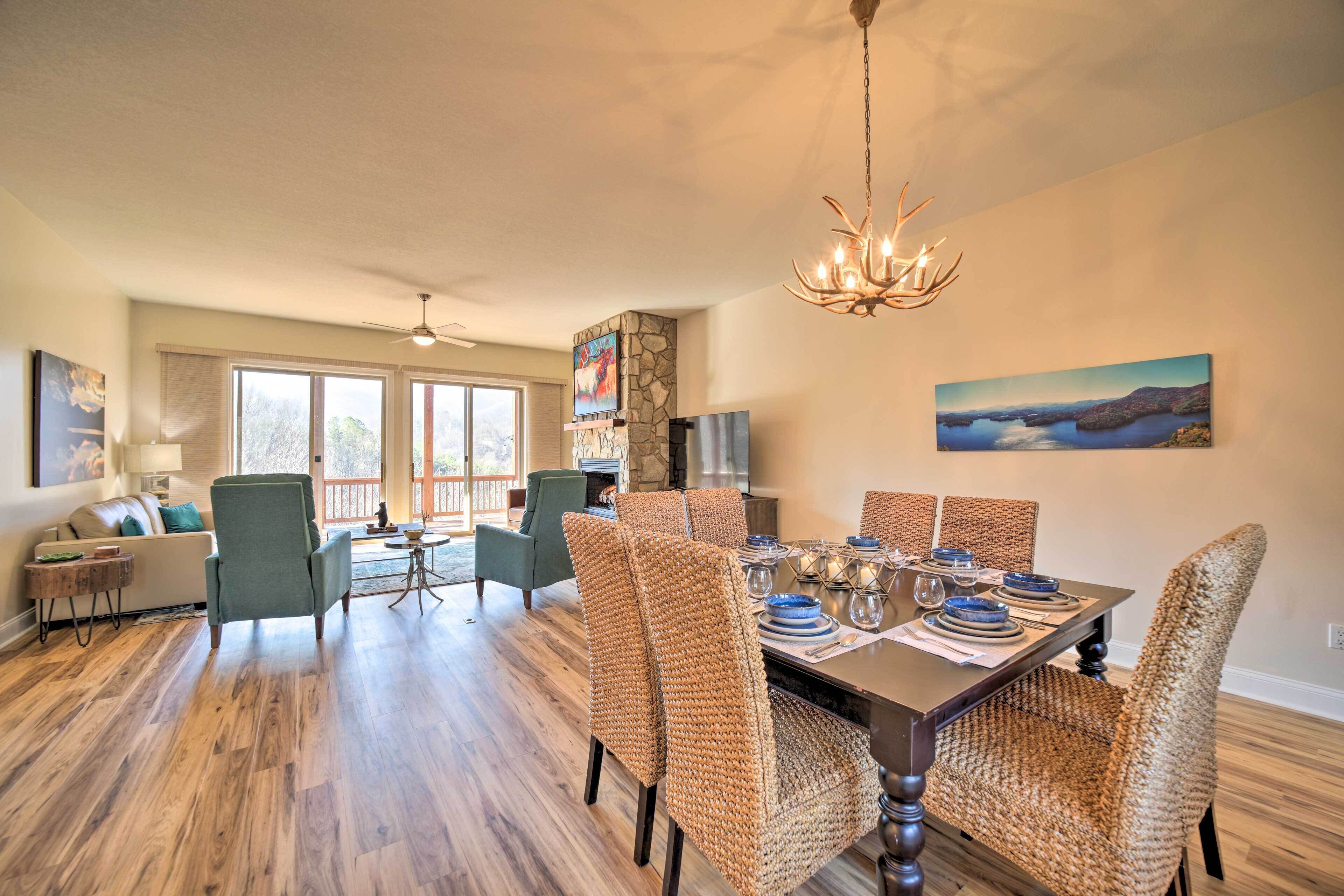 Un coin repas avec une table en bois dressée pour six personnes, entourée de chaises en osier, d’un lustre et d’un tableau accroché au mur.