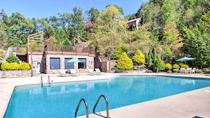 A rectangular swimming pool with clear blue water, surrounded by a stone deck. There are lounge chairs and umbrellas, and a wooden deck with a railing. - Beautiful Whittier Condo w/ Deck + Mtn Views! (Whittier)