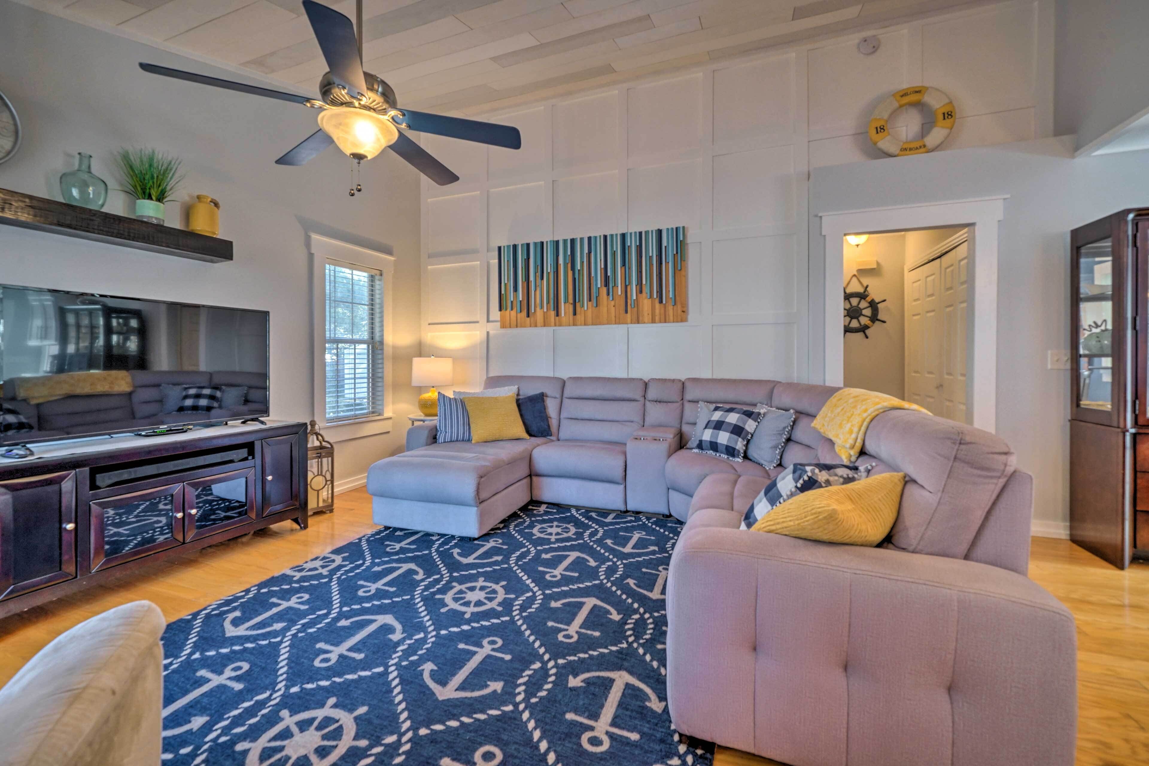 A modern living room with a sectional sofa, a ceiling fan, a flat-screen TV, and a decorative rug with nautical patterns.