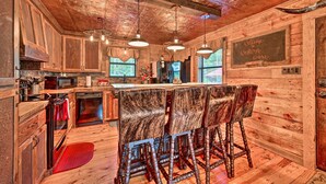 A rustic kitchen with wooden cabinets, a bar area with stools, and a chalkboard sign. - 2 Mi to Choctaw Landing: 'western Daze' Cabin! (Broken Bow)