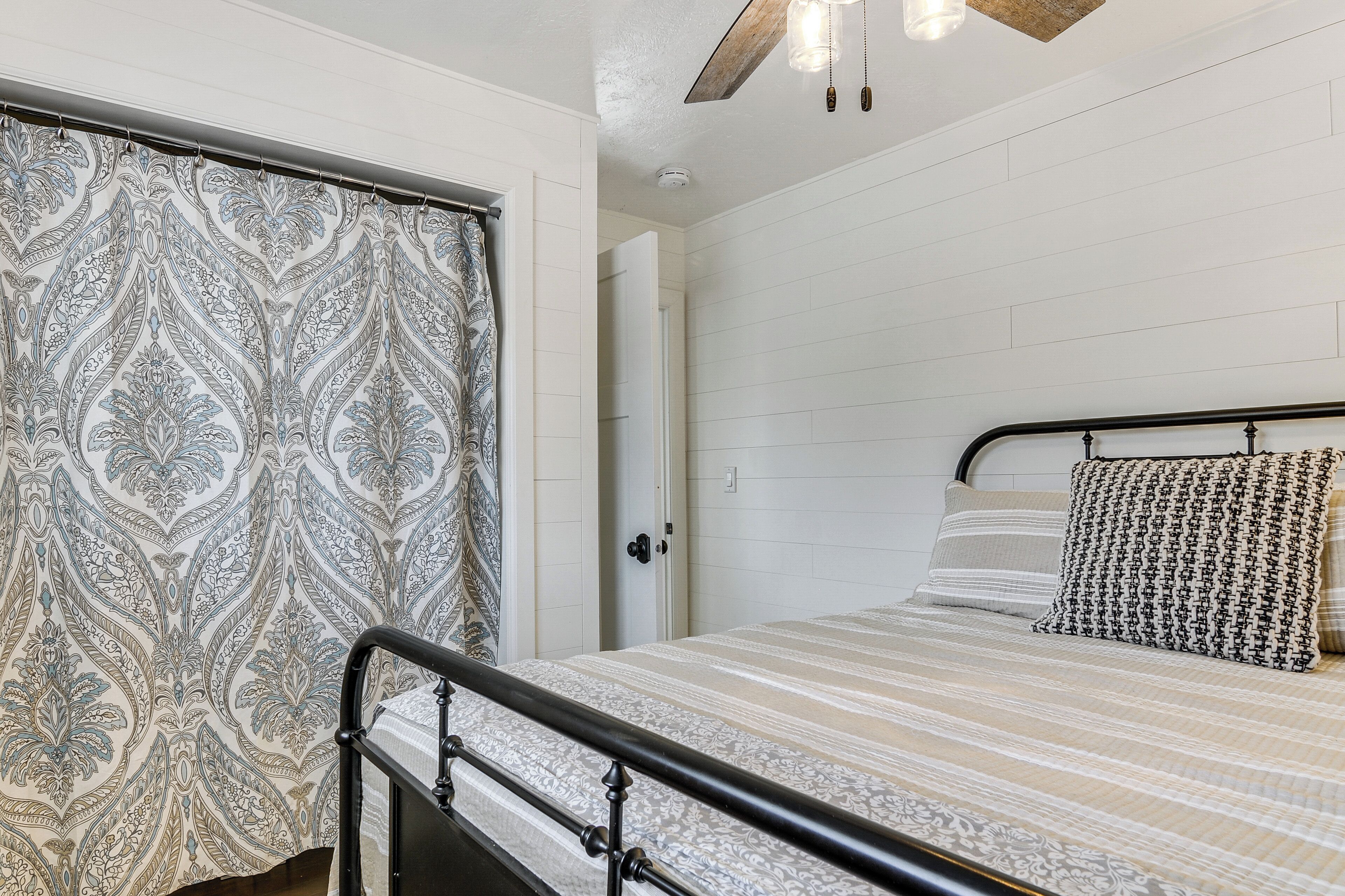 A bedroom with a bed, a patterned curtain, a ceiling fan, and a door leading to another room.