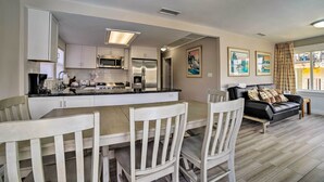 A modern kitchen with white cabinets, stainless steel appliances, and a central island. There is a dining area with white chairs and a black leather sofa. The room has a neutral color palette and features artwork on the walls. - Ocean-view Home - Walk to Avalon City Center! (Avalon)