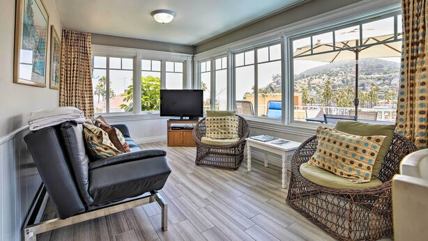 A sunroom with a sofa, two wicker chairs, a TV, and a view of a hilly landscape. - Ocean-view Home - Walk to Avalon City Center! (Avalon)