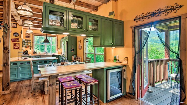 Une cuisine avec des meubles de couleur verte, un îlot central en bois doté de tabourets et une vue dégagée sur l’extérieur grâce à une grande fenêtre.