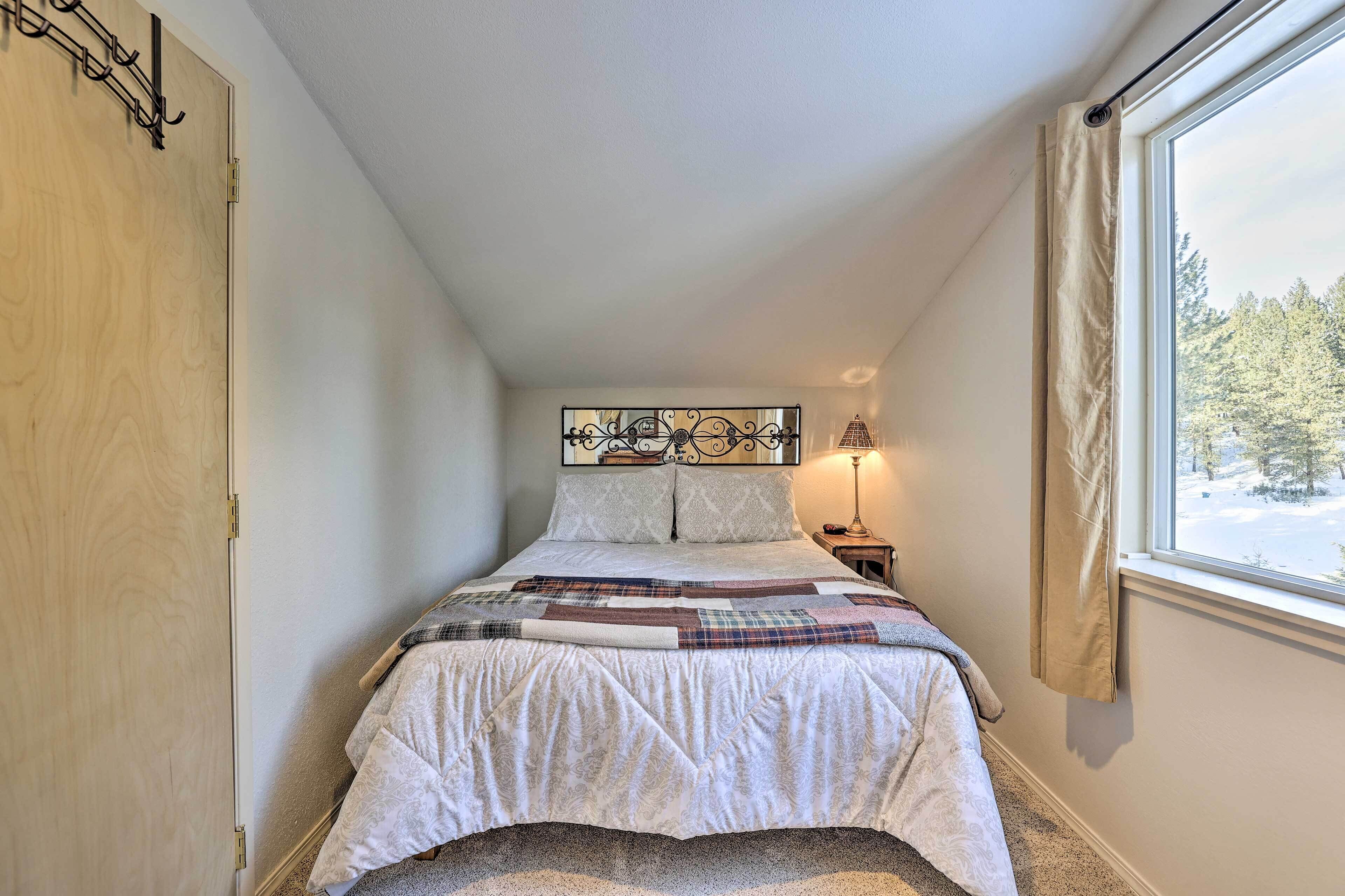 A bedroom with a bed, a window with a view of trees, a nightstand with a lamp, and a door.