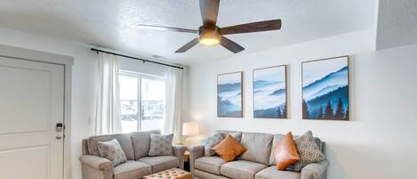 A living room with two grey sofas, a coffee table, a ceiling fan, and three framed landscape paintings on the wall.