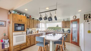 A kitchen with wooden cabinets, a microwave, a stove, a hanging pot rack, and a central island with bar stools. - Charming Sequim House w/ Olympic Peninsula Views! (Sequim)