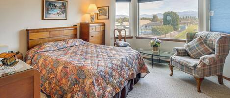 A bedroom with a large bed, a chair, a dresser, a lamp, and a view of the outdoors.
