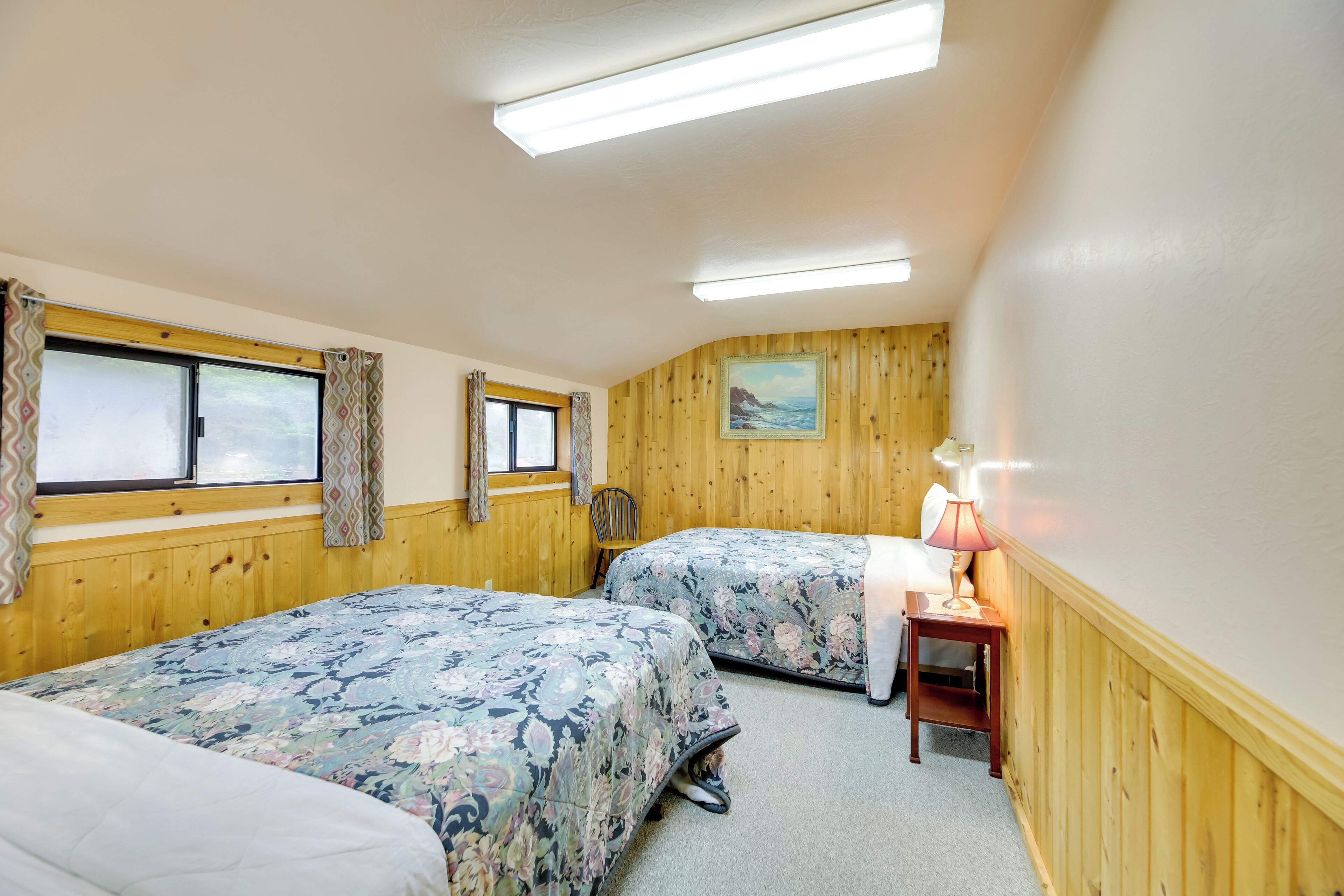 A room with two beds, wooden walls, a painting, a lamp, and a chair.