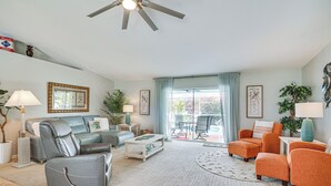 Een ruime woonkamer met een grijze leren bank, een oranje fauteuil en een witte salontafel. Er is een plafondventilator, een groot raam met gordijnen en uitzicht op de omgeving.