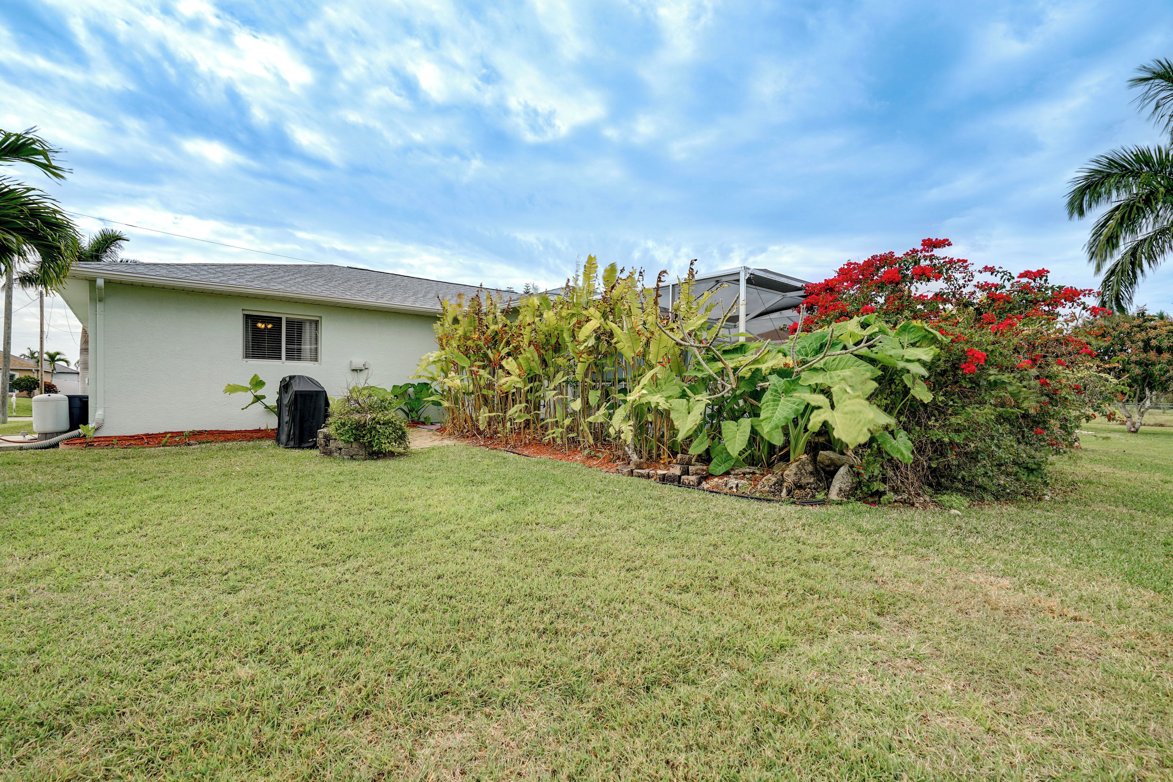 A single-story house with a well-maintained lawn, a small garden with red flowers, and a palm tree to the left.