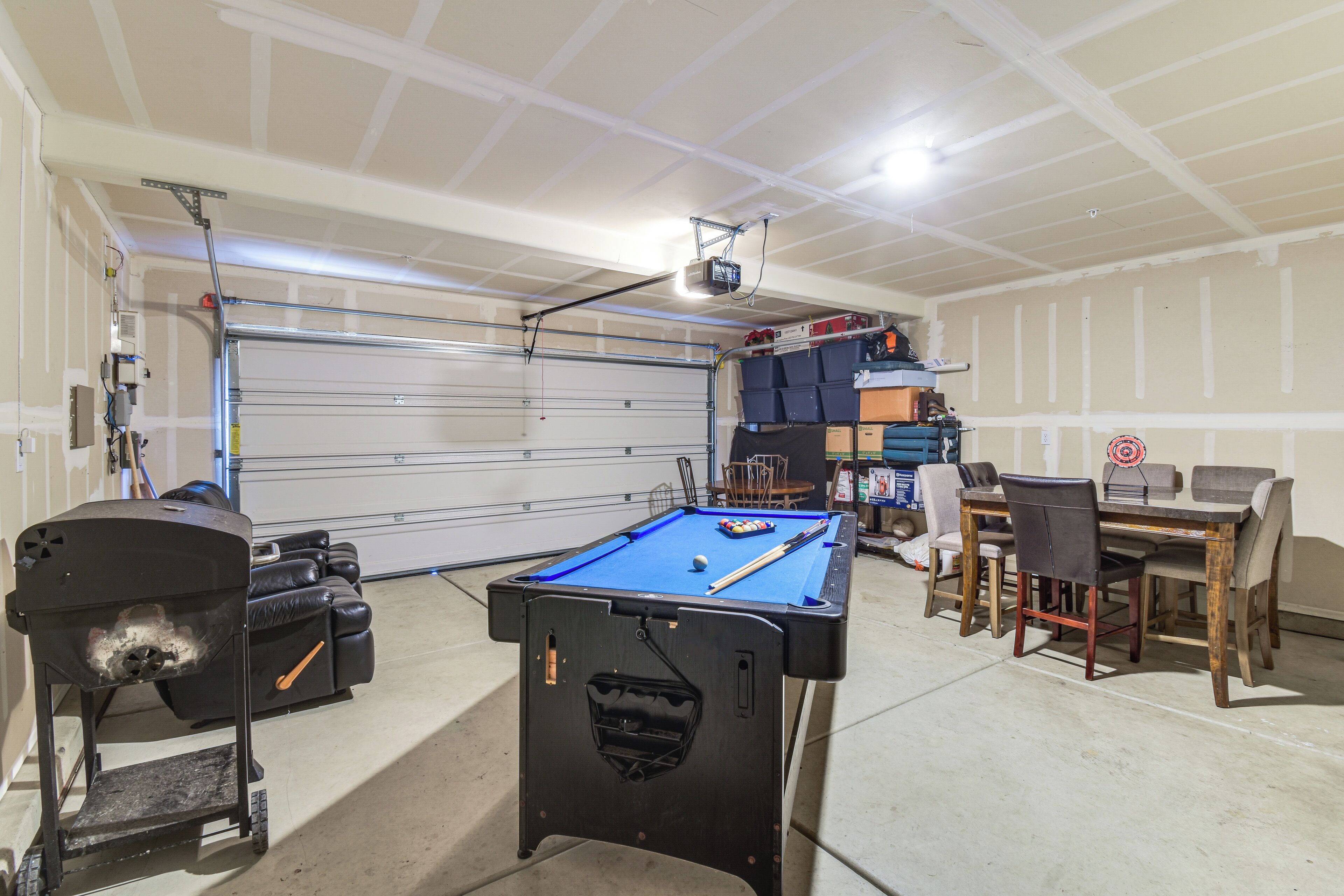 A pool table in a garage with a ceiling-mounted projector, a dining table with chairs, and storage shelves.