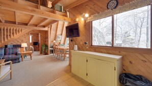 L’intérieur d’un chalet accueillant, avec un plafond en bois, un téléviseur fixé au mur, un coin repas et une fenêtre donnant sur la forêt.