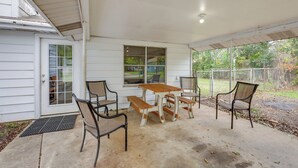 Un porche techado con una mesa y bancos de madera, una puerta con ventana y vista a un patio cercado.