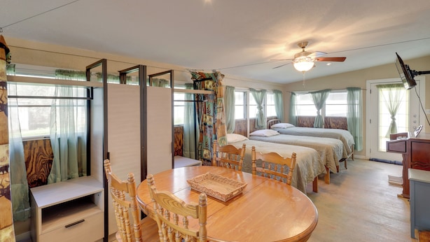 Una habitación con una mesa de comedor de madera, dos camas, un ventilador de techo y una ventana con cortinas.