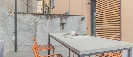Ein kleiner Sitzbereich im Freien mit Tisch und zwei Stühlen, eine Betonwand und eine Holztür mit Fensterläden.