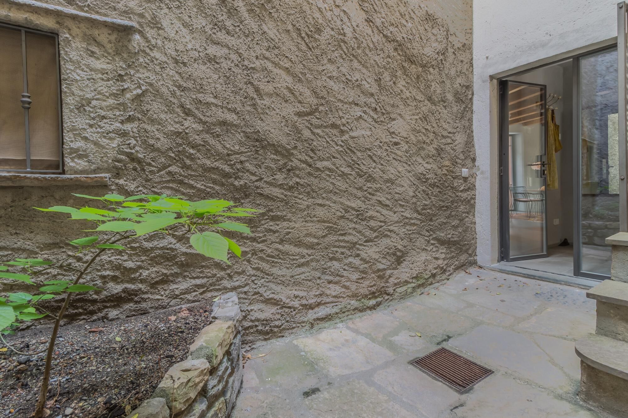 A stone exterior wall with a small plant, a window with a metal grill, and a glass door leading inside.