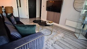 A modern living room with a dark blue sofa, a round coffee table, and a flat-screen TV mounted on the wall. - 1 Bed Flat - Sleeps 2 - Jacuzzi - Parking - Wifi (Birmingham)