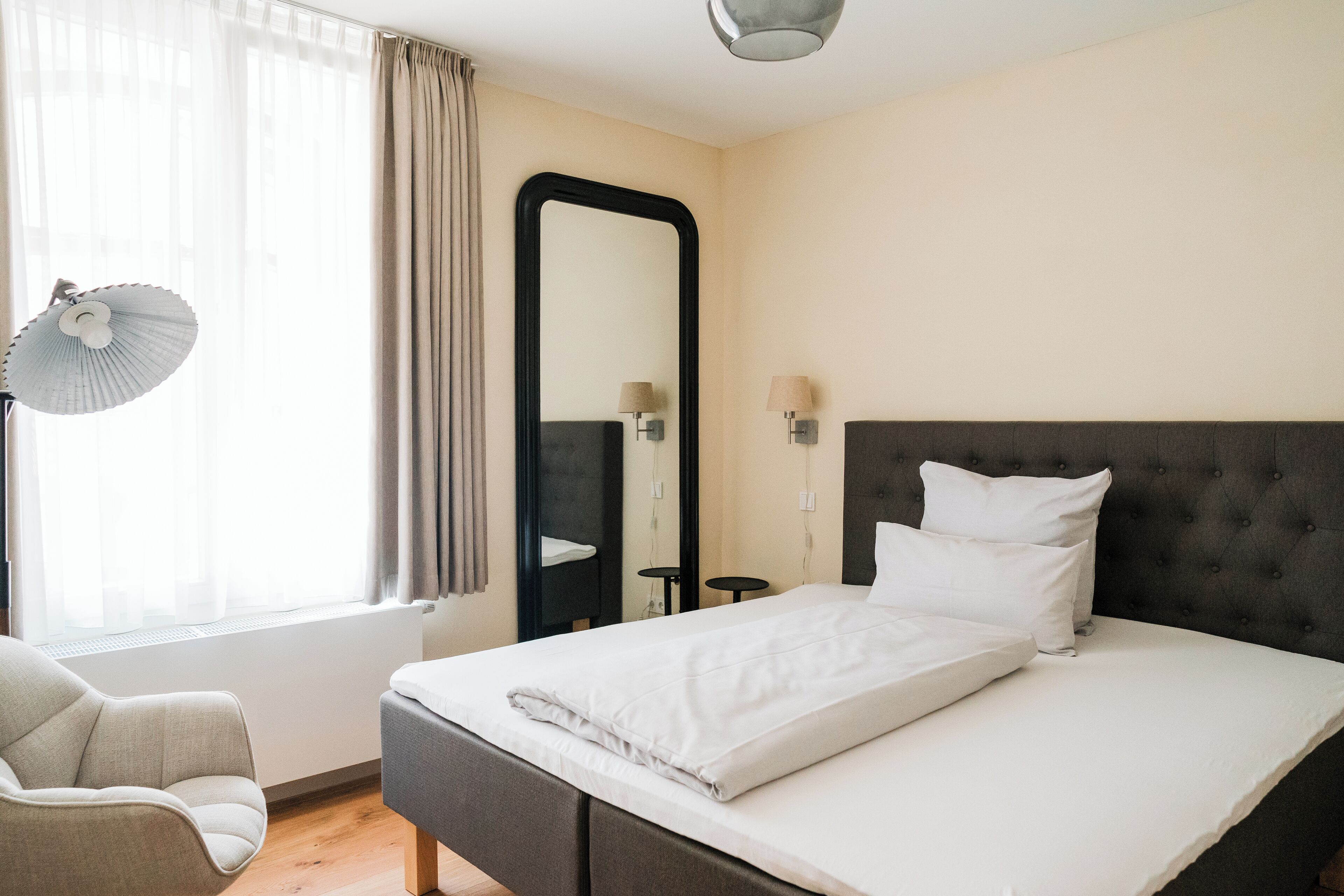 A hotel room with a bed, a chair, a mirror, and a window with curtains.