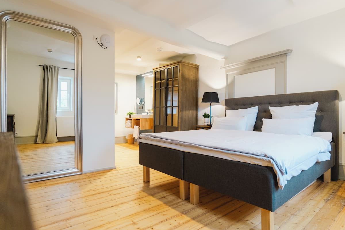 Une chambre à coucher avec un grand lit, un plancher en bois, une armoire, un miroir et une table de chevet avec une lampe.