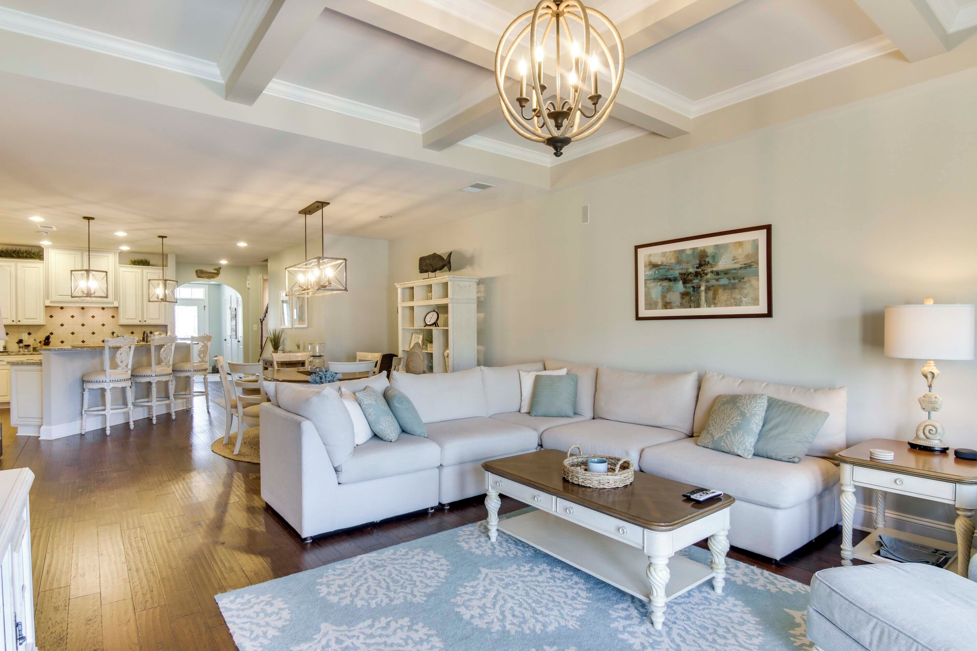 A spacious living room with a white sectional sofa, a wooden coffee table, and a chandelier. A kitchen with white cabinets and a dining area with a table and chairs are visible in the background.