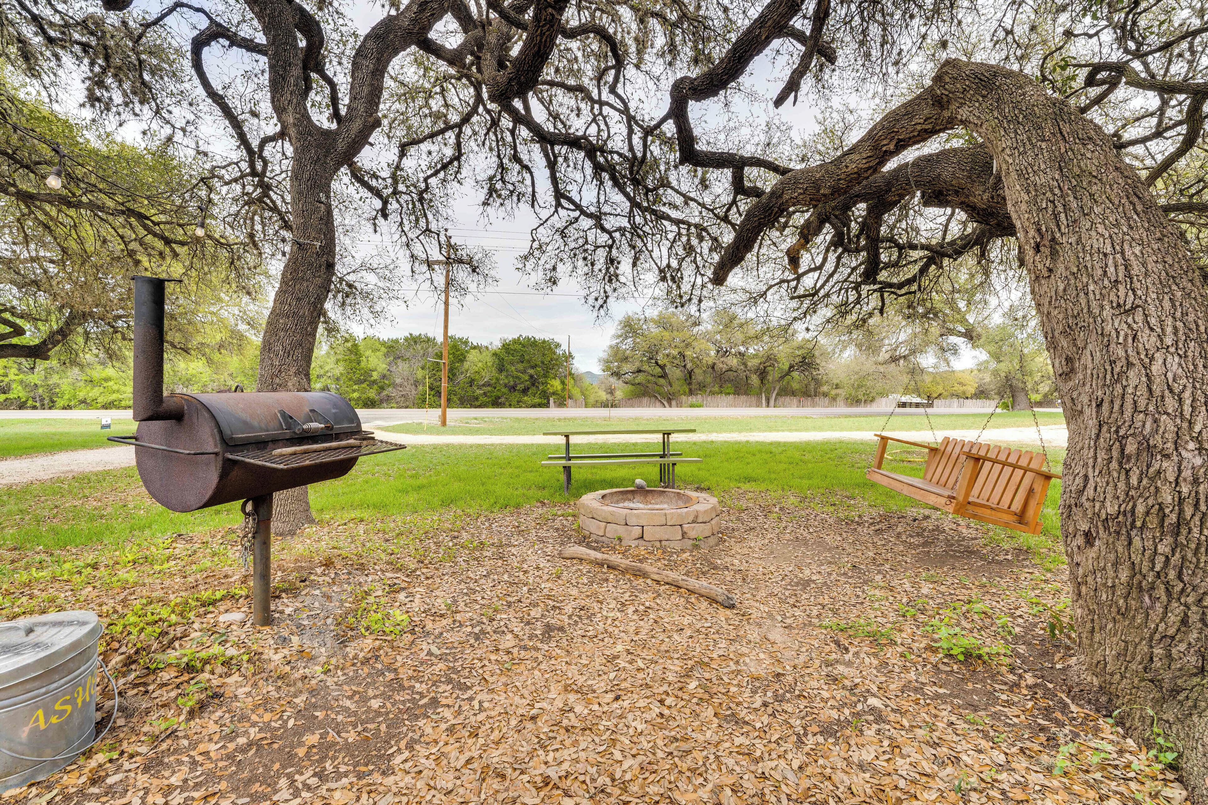 A barbecue area with a grill, a fire pit, a picnic table, and a swing under a large tree.
