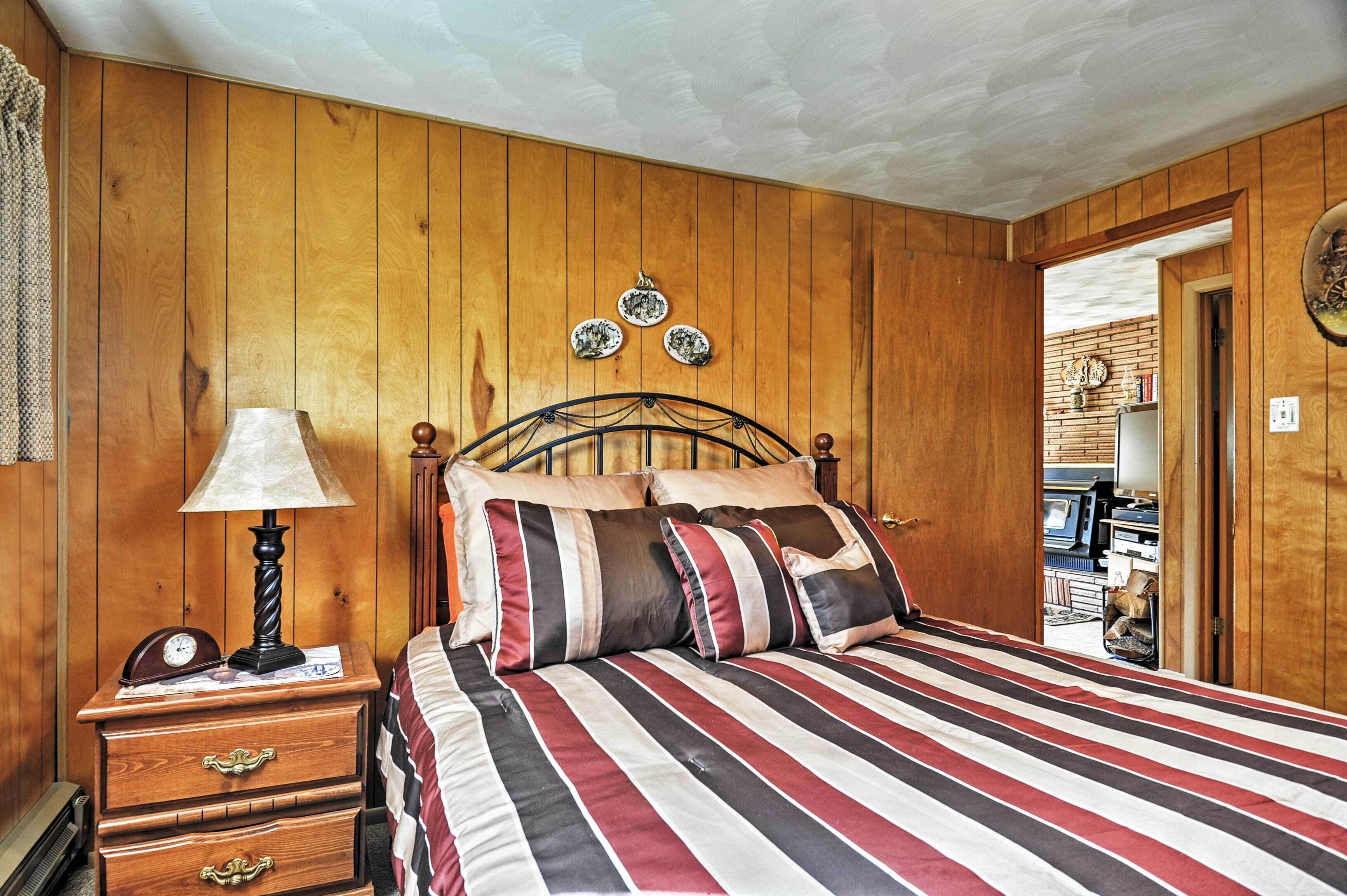A wooden-paneled bedroom with a bed, a nightstand with a lamp, and a door leading to another room.